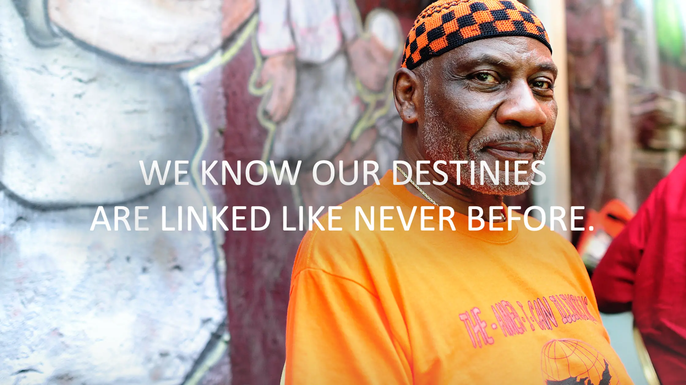an african american man standing near a wall covered in grafitti with the text "WE KNOW OUR DESTINIES ARE LINKED LIKE NEVER BEFORE."